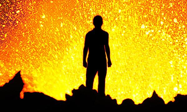 silhouette of person standing in front of a volcanic eruption. Yellow lava flares in front of him.