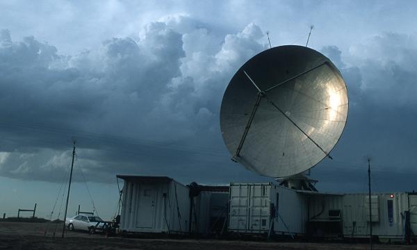 Image of a large ground radar placed above large metal containers. Clouds in the background indicate a looming storm. 