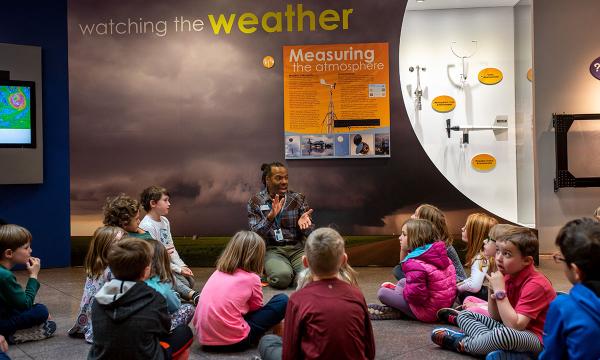 Tim Barnes kneels on the floor in front of a weather exhibit with a group of children sitting around him